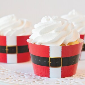 Może przedstawiać: Trzy czerwono-białe foremki do muffinek z czarnym paskiem i złotą klamrą. Foremki są ozdobione tak, aby przypominały pasek Świętego Mikołaja. Muffinki są przyozdobione białym kremem.