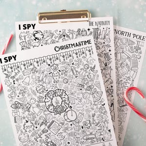 May include: A clipboard holds three black and white "I Spy" activity sheets with Christmas themes. The sheets are titled "Christmastime", "The Nativity", and "North Pole". Red and white candy canes are placed around the sheets.