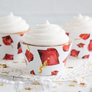May include: Three white cupcakes with white frosting, decorated with red graduation caps on the cupcake wrappers. The cupcakes are on a white doily with gold confetti.