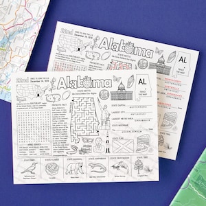 Op de afbeelding: Twee witte activiteitenvellen met het thema Alabama, met illustraties, een doolhof en een woordzoeker. De vellen bevatten het staatsmotto, de hoofdstad en de bijnaam, met een kaart en andere staatssymbolen. De vellen liggen op een blauw oppervlak.