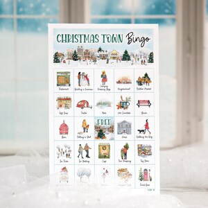 May include: A Christmas Town Bingo card with a watercolor illustration of a winter town scene. The card has 25 squares with different images, including a snowman, a Christmas tree, a gift shop, a hot chocolate stand, and a walking dog. The free space is in the center of the card.