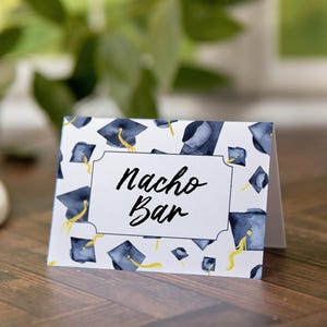 May include: White card with a blue and gold graduation cap pattern. The card has a white rectangle with a black border and the text "Nacho Bar" in black cursive writing.