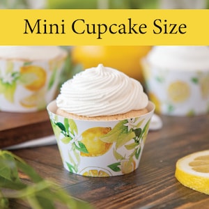 May include: Mini lemon cupcake with white frosting in a white cupcake liner with a lemon and leaf pattern. The cupcake is on a wooden table with a lemon slice next to it.