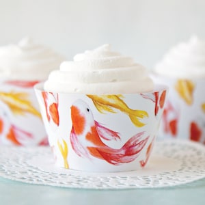 May include: Three cupcakes with white frosting in decorative paper cups. The cups are white with a repeating pattern of orange, yellow, and red koi fish. The cupcakes sit on a white, lacy paper doily.