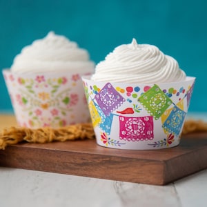 May include: Two cupcakes with white frosting, one in a colorful floral cupcake liner and the other in a cupcake liner with a colorful Mexican paper cutout design.