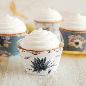 May include: Four cupcakes with white frosting sit in decorative paper liners. The liners feature a floral and succulent design in shades of blue, gray, and white. The cupcakes are on a light wooden surface.