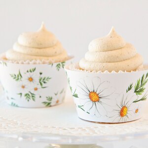 May include: Two white cupcakes with vanilla frosting and a daisy pattern on the cupcake liners.