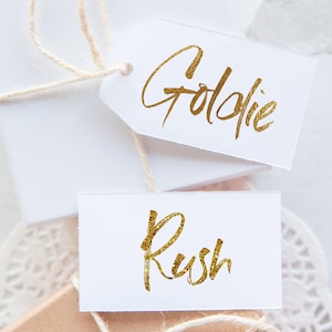 May include: Two white gift tags with gold glitter lettering. The top tag says "Goldie" and the bottom tag says "Rush".