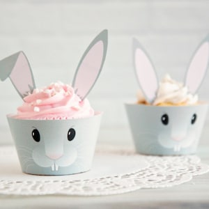 May include: Two cupcakes decorated with bunny ears and faces. The cupcakes are frosted with pink and white frosting and have gray bunny ears and faces.