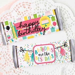 Pode incluir: Dois embrulhos de barras de chocolate com designs de aniversário. O embrulho superior é rosa e branco com o texto "feliz aniversário" e ícones coloridos de festa de aniversário. O embrulho inferior é branco com um padrão de confete colorido e o texto "ANIVERSÁRIOS são os MELHORES DIAS" em uma faixa.