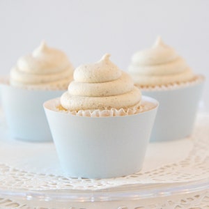 May include: Three cupcakes with light blue paper cups and white frosting. The frosting is swirled and has a light brown speckled appearance. The cupcakes are on a clear, decorative plate.