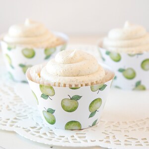 Op de afbeelding: Drie cupcakes met witte frosting en vanilleboonvlekken in papieren vormpjes. De vormpjes zijn versierd met groene aquarelappels op een witte achtergrond. De cupcakes staan op een witte, kanten onderlegger.