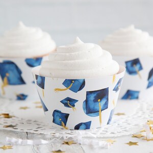 May include: Three white cupcakes with blue and gold graduation cap cupcake wrappers. The cupcakes are on a white doily with gold confetti.