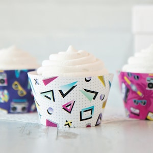 Puede incluir: Tres cupcakes con glaseado blanco y envoltorios de cupcakes con patrones geométricos coloridos. Los envoltorios presentan una mezcla de formas geométricas rosas, azules, moradas y amarillas.