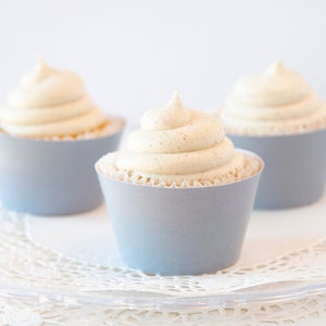 May include: Three cupcakes with light blue paper liners and white frosting. The cupcakes are arranged on a clear, decorative plate with a white doily underneath. The frosting has a swirl design and a light brown color.
