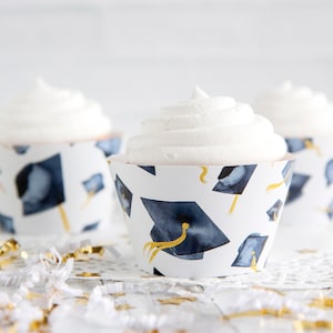 May include: Three white cupcakes with white frosting and blue and gold graduation cap cupcake wrappers. The cupcakes are on a white surface with gold and white confetti.