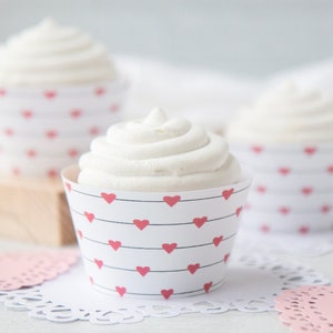 Puede incluir: Tres cupcakes con glaseado blanco en moldes de papel blancos decorados con corazones rojos. Los cupcakes están sobre blondas blancas y un mantelito de papel rosa claro. El fondo está borroso.