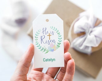 Puede incluir: Una etiqueta de regalo blanca con una corona verde, una cruz dorada y el texto "He is Risen". La etiqueta también tiene el nombre "Catelyn" escrito en verde.