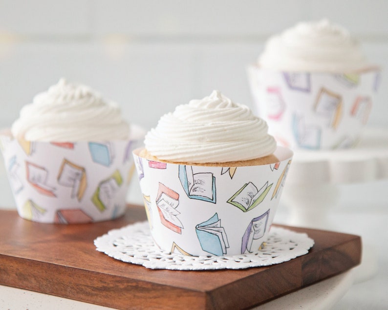 May include: Three white cupcakes with vanilla frosting, decorated with cupcake wrappers featuring a colorful book pattern. The wrappers have a white background with illustrations of open books in various colors, including blue, pink, yellow, and green.