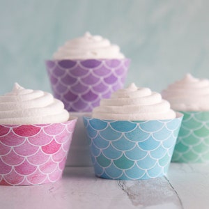 May include: Four cupcakes with white frosting in mermaid scale patterned paper cases. The cases are in pink, blue, purple, and green. The cupcakes are arranged on a light blue surface.
