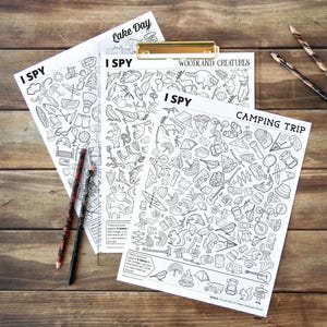 May include: Three black and white "I SPY" activity sheets with different themes, including "Lake Day", "Woodland Creatures", and "Camping Trip". Two pencils rest on the wooden surface. The sheets are on a wooden clipboard.