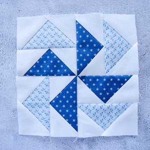 May include: A square quilt block featuring a pinwheel design. The block is composed of white fabric and triangles of blue fabric. Some blue triangles have a white star pattern, while others have a delicate floral pattern.