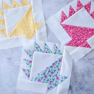 May include: Three colorful quilt blocks with white, yellow, pink, and blue floral patterns. Each block features a unique geometric design, showcasing a basket or sunburst motif. The blocks are set against a light gray background.