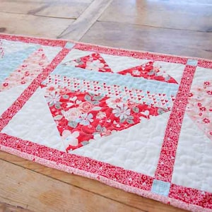 Könnte beinhalten: Gesteppter Tischläufer mit Herzmuster. Das Herz besteht aus rotem Blumenstoff, umgeben von weißen und hellblauen Abschnitten. Der Läufer ist mit einem roten Blumenmuster eingefasst und misst ca. 30 cm breit und 76 cm lang.