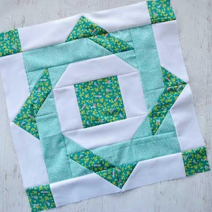 May include: A square quilt block with a geometric design. The block features white, teal, and green patterned fabric. The center square is green with a floral pattern, surrounded by white and teal shapes. The block is approximately 10 inches square.