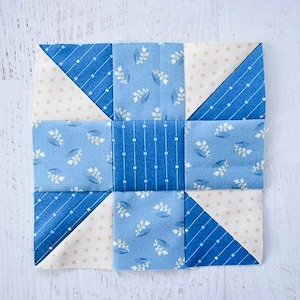 May include: A square quilt block featuring a starburst design. The block is composed of various blue fabrics, including floral prints, striped patterns, and solid blue. Cream-colored fabric with small dots is also used. The block is likely part of a larger quilt.