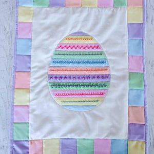 May include: A pastel-colored quilted wall hanging with an embroidered Easter egg design. The egg is surrounded by a border of squares in various shades of pink, yellow, green, and purple.