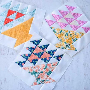 May include: Three colorful quilt blocks with white borders. Each block features a different pattern: red and blue triangles, pink triangles with yellow floral, and blue triangles with orange floral. The blocks are arranged on a white surface.