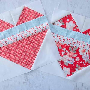 May include: Two heart-shaped quilt blocks. The hearts are made of red fabric with white and pink patterns, and floral designs. A light blue strip and a patterned strip with tiny hearts cross the center of each heart. The background is white.