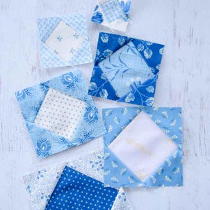 Puede incluir: Varios cuadrados de tela de patchwork azules y blancos con diversos patrones, incluyendo florales y lunares, están dispuestos sobre una superficie de madera blanca. Los cuadrados son en tonos de azul claro y oscuro, con un cuadrado central blanco.