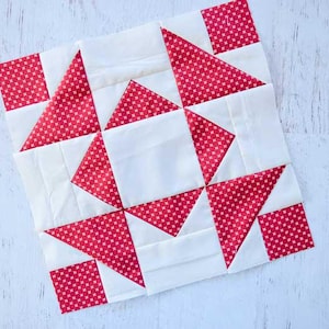 Puede incluir: Un bloque de edredón cuadrado con un diseño geométrico en rojo y blanco. La tela roja tiene un pequeño estampado de lunares. El bloque está compuesto por cuadrados y triángulos, creando un patrón en forma de estrella.