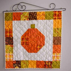 May include: A quilted wall hanging featuring a large orange pumpkin design. The pumpkin is centered on a white background with a diamond pattern. The border is made of colorful patchwork squares in fall colors, with a decorative metal hanger.