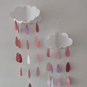 May include: Two white paper clouds with pink and purple paper raindrops hanging from them. The clouds are hanging from the ceiling.