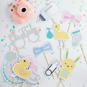 Op de afbeelding: Een set van 10 fotobooth-rekwisieten voor een babyshower met verschillende schattige ontwerpen, waaronder een babyfles, een strik, een ooievaar, een sok, een strikje, een eend, een speen, een veiligheidsspeld, een konijn en een bril met de tekst "Oh Baby".