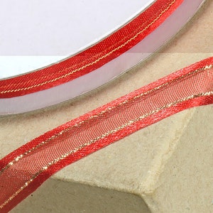Könnte beinhalten: Rotes, durchsichtiges Band mit goldfarbenem Metallic-Besatz. Das Band ist auf einer weißen Spule aufgerollt.