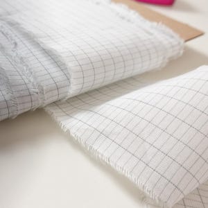 Könnte beinhalten: Eine Rolle weißen Stoffes mit grauem Gittermuster, teilweise abgerollt, auf einer braunen Kartonbasis. Der Stoff hat einen ausgefransten Rand, was auf ein Bastel- oder Textilprojekt hindeutet.