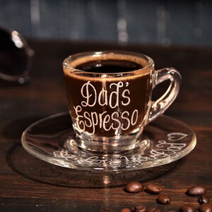 May include: A clear glass espresso cup with the words "Dad's Espresso" etched on the side. The cup is filled with dark coffee and sits on a clear glass saucer. Coffee beans are scattered on a dark wooden surface.