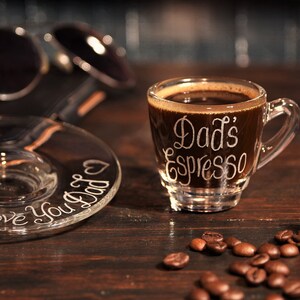 May include: A clear glass espresso cup and matching saucer set. The cup features the white etched text "Dad's Espresso" and the saucer is etched with the text "Love You Dad" accompanied by two small heart icons, filled with dark coffee.