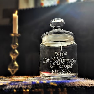 May include: A clear glass jar with a lid, engraved with a cross and the text "On your First Holy Communion Isla McDonald 6/5/2023". The jar is sitting on a colourful rug.