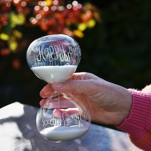 May include: Clear glass hourglass with white sand, held in a hand. The top of the hourglass reads "Happy Birthday" in cursive script. The bottom reads "XX Cool Grandma". The background is blurred with fall colors.