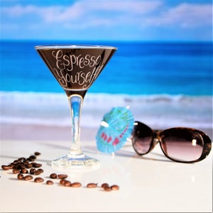 A classic espresso martini shaped cocktail glass rests on a table near the beach. The glass has been hand engraved in a calligraphy font with the words Espresso Yourself! and a date and message engraved on the base of the glass