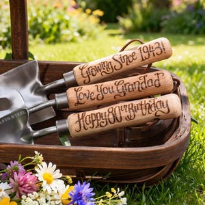 a set of three hand gardening tools with stainless steel heads and wooden handles, a trowel, a fork and a planter. the wooden handles have been pyrographed in a calligraphy font with the messages 'Growing since 1943', 'Love You Grandad' and 'Happy 80th Birthday'. The tools are in a natural trug on a lawn in a sunny garden with wild meadow flowers in the foreground