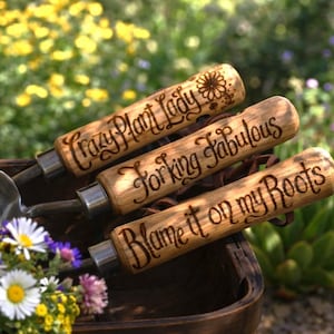 May include: Three metal garden trowels with light brown wooden handles. Each handle features dark wood-burned text: Crazy Plant Lady, Forking Fabulous, and Blame it on my Roots. The tools are arranged together with a small bouquet of daisies.