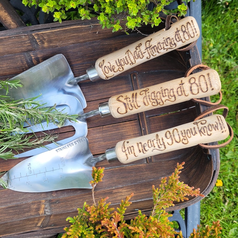 May include: Three stainless steel garden tools with wooden handles. The handles are engraved with phrases like "Love you Dad - Amazing at 80!" and "Still Jigging at 80!". The tools are arranged in a wooden basket with greenery.