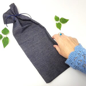 May include: A dark gray wine bottle bag made of fabric, tied at the top with a navy blue string. The bag is on a white surface with green leaf decorations.