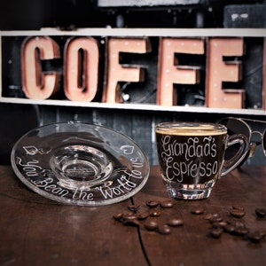 May include: A clear glass espresso cup and matching saucer set. The cup features the white etched text "Grandad's Espresso" and the saucer features the white etched text "You Bean the World to us" with small heart icons. Scattered roasted coffee beans.